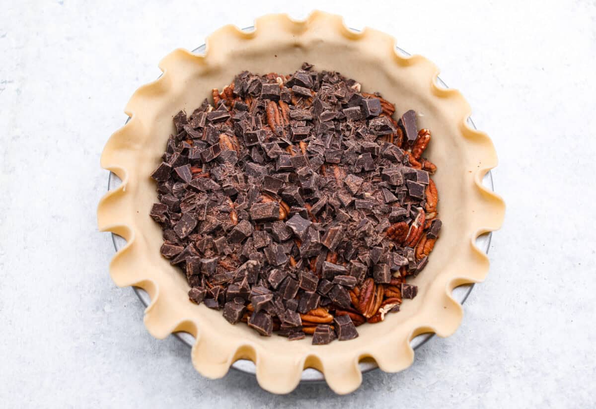 Chocolate pecan pie is a sweet, decadent dessert with a buttery crust filled with gooey caramel, crunchy pecans, and rich chocolate. It's the perfect pie to share this Thanksgiving. It will knock your family's socks off! Overhead shot of a layer of chocolate chunks over the pecans in a pie crust.