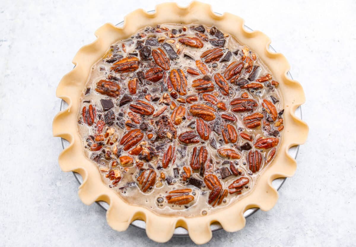 Chocolate pecan pie is a sweet, decadent dessert with a buttery crust filled with gooey caramel, crunchy pecans, and rich chocolate. It's the perfect pie to share this Thanksgiving. It will knock your family's socks off! Overhead shot of the pie crust with pecans, chocolate and filling inside ready to go in the oven.