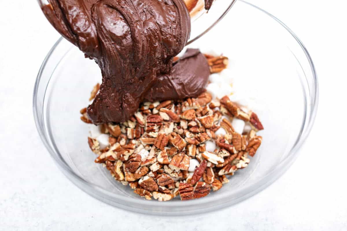 These decadent rocky road bars are the best no-bake treat that you can whip up in no time when that sweet tooth hits. Fudgy chocolate, soft marshmallows, and chewy pecans combine to make the most amazing dessert. Angle shot of someone pouring the chocolate mixture into the nuts and marshmallows.