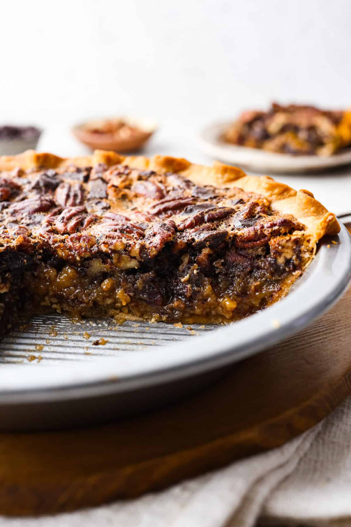 Chocolate pecan pie is a sweet, decadent dessert with a buttery crust filled with gooey caramel, crunchy pecans, and rich chocolate. It's the perfect pie to share this Thanksgiving. It will knock your family's socks off! Side shot of chocolate pecan pie in a pie pan with a slice taken out showing the gooey inside.