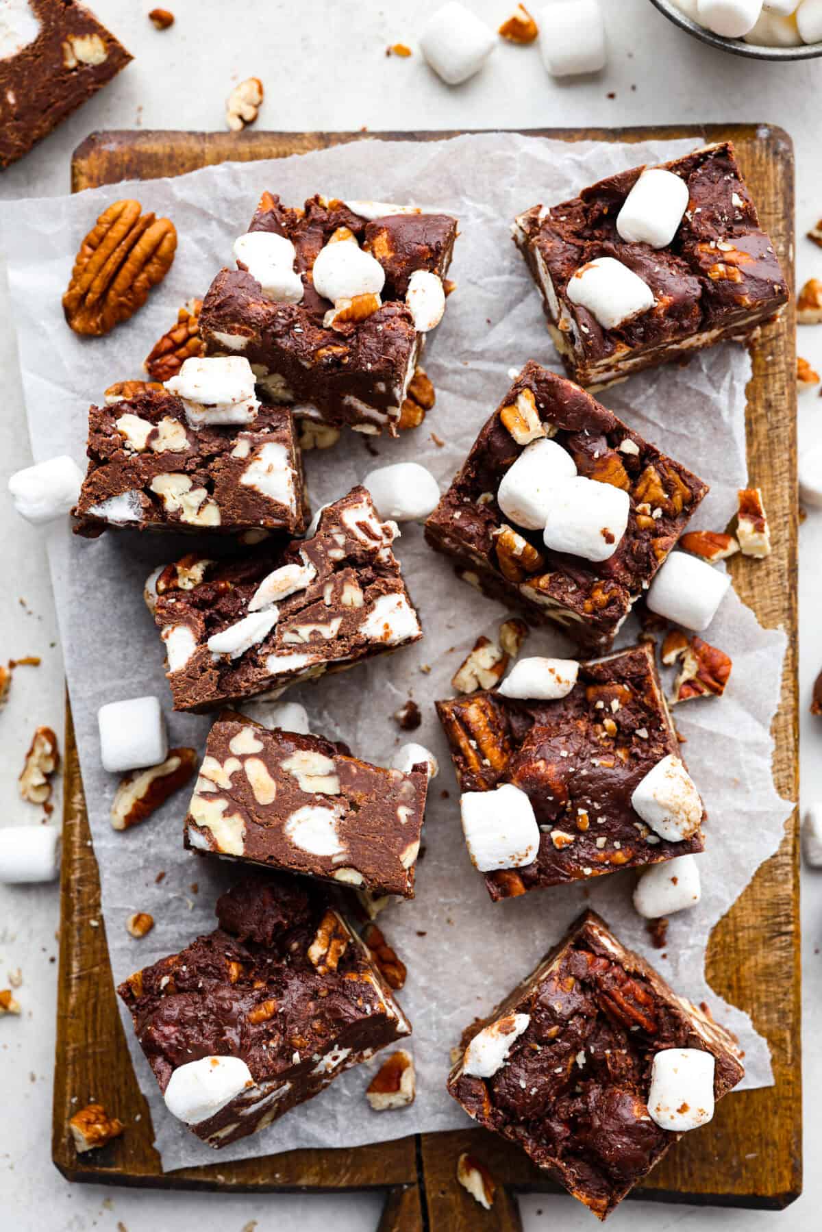 These decadent rocky road bars are the best no-bake treat that you can whip up in no time when that sweet tooth hits. Fudgy chocolate, soft marshmallows, and chewy pecans combine to make the most amazing dessert. Overhead shot of rocky road bars cut up on parchment paper on a wooden cutting board.