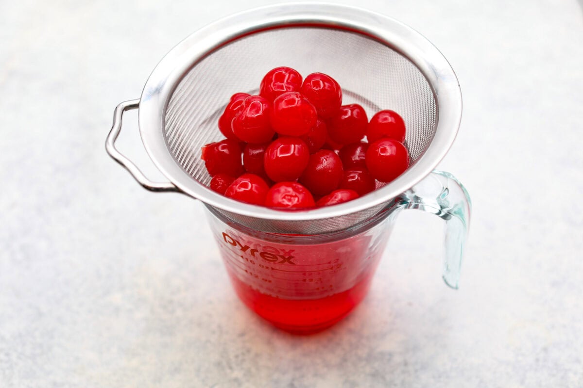 Homemade cherry cordials are a tasty, nostalgic treat! They're made with sweet cherries that are coated in a rich chocolate shell. Make them for Christmas or Valentine’s Day, and watch them disappear! Angle shot of cherries in a strainer over a measureing cup.