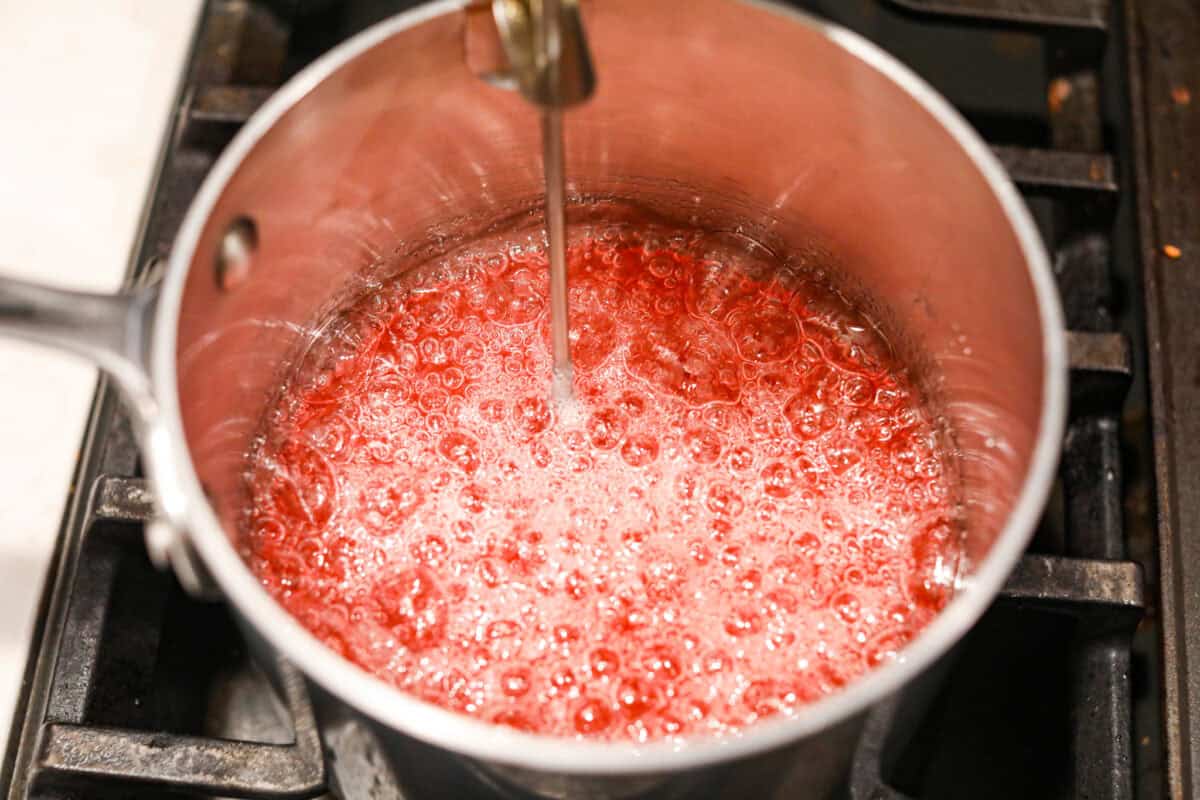 Homemade cherry cordials are a tasty, nostalgic treat! They're made with sweet cherries that are coated in a rich chocolate shell. Make them for Christmas or Valentine’s Day, and watch them disappear! Angle shot of sugars boiling in the saucepan with a candy thermometer sticking out.