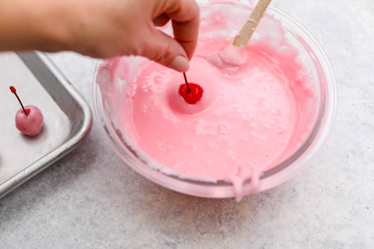 Homemade cherry cordials are a tasty, nostalgic treat! They're made with sweet cherries that are coated in a rich chocolate shell. Make them for Christmas or Valentine’s Day, and watch them disappear! Angel shot of someone dipping cherries into the fondant.
