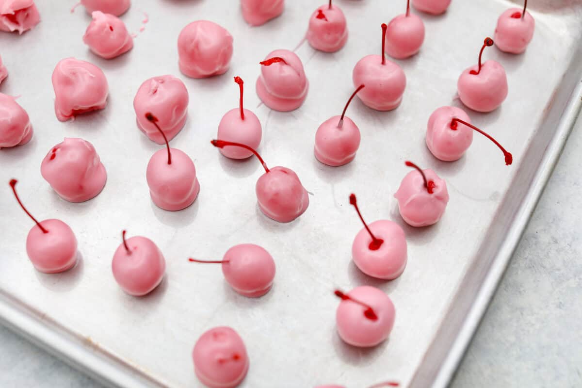 Homemade cherry cordials are a tasty, nostalgic treat! They're made with sweet cherries that are coated in a rich chocolate shell. Make them for Christmas or Valentine’s Day, and watch them disappear! Overhead shot of fondant dipped cherries on a baking sheet.