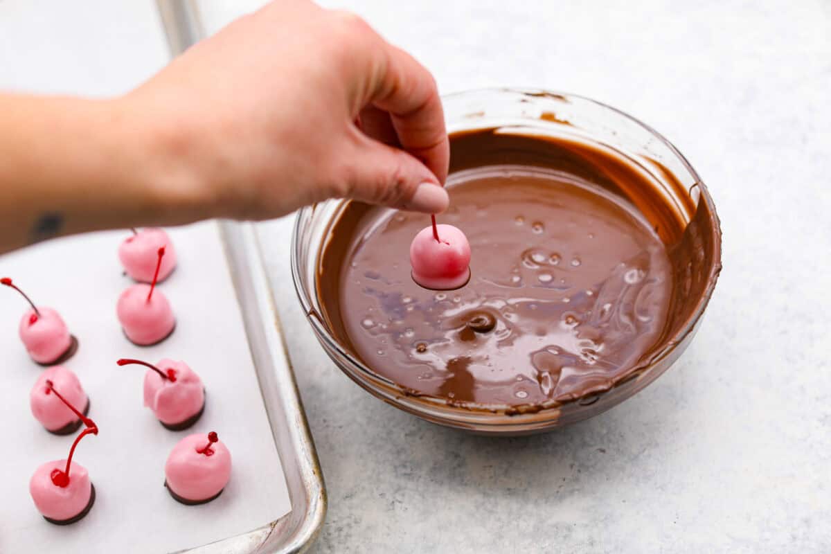 Homemade cherry cordials are a tasty, nostalgic treat! They're made with sweet cherries that are coated in a rich chocolate shell. Make them for Christmas or Valentine’s Day, and watch them disappear! Angle shot of someone dipping fondant dipped cherries into the chocolate.