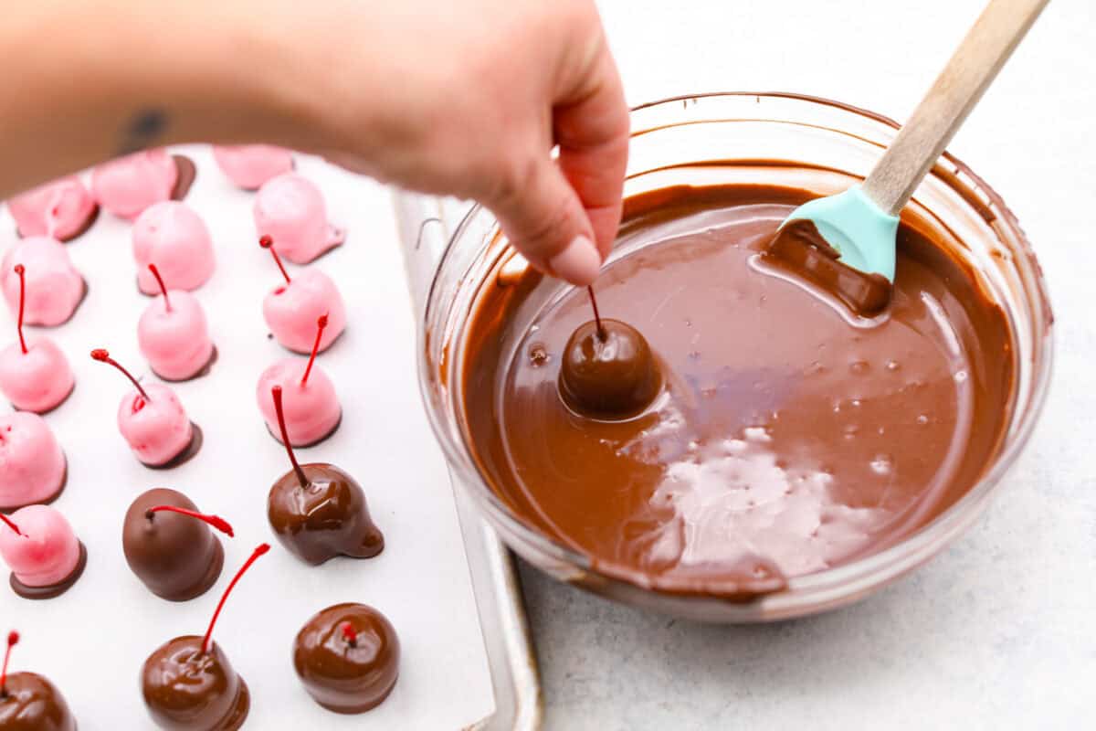 Homemade cherry cordials are a tasty, nostalgic treat! They're made with sweet cherries that are coated in a rich chocolate shell. Make them for Christmas or Valentine’s Day, and watch them disappear! Angle shot of someone holding a chocolate dipped cherry over the bowl of chocolate.