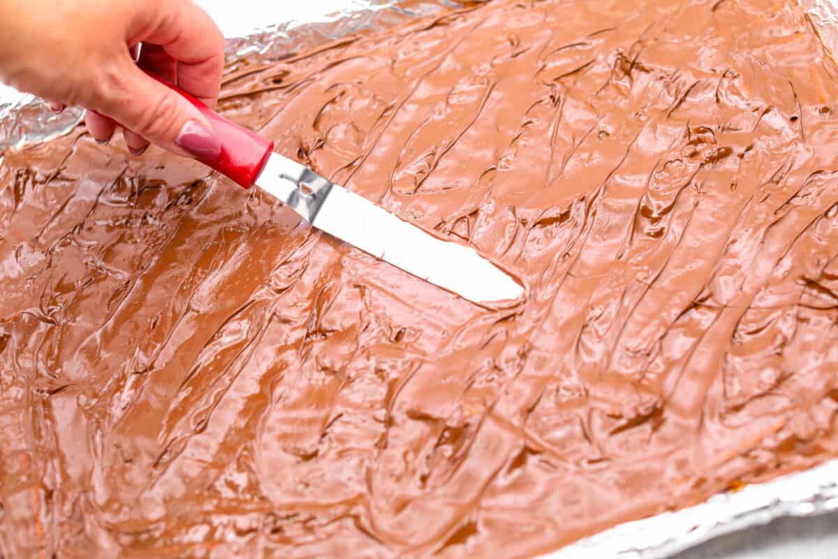 Pretzel Christmas Crack is the best holiday treat! This addictive combination of salty pretzels, rich toffee, and chocolate is sure to be a hit at any party or gathering. Angle shot of someone spreading the melted chocolate chips over the caramel mixture.