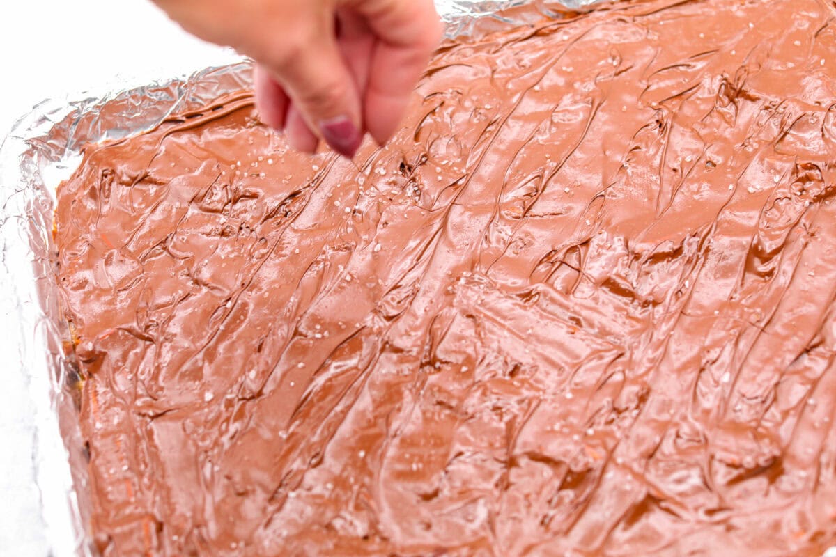 Pretzel Christmas Crack is the best holiday treat! This addictive combination of salty pretzels, rich toffee, and chocolate is sure to be a hit at any party or gathering. Angle shot of someone sprinkling flaky sea salt over the chocolate.