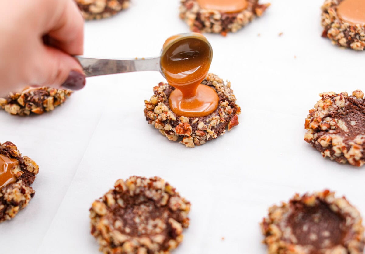 Turtle Cookies are a baked, bite-size brownie loaded with pecans, then filled with a creamy caramel center for a rich, decadent holiday cookie! These turtle cookies are always a huge hit and are gone in minutes Angle shot of someone pouring the melted caramel into the indents in the baked cookies.