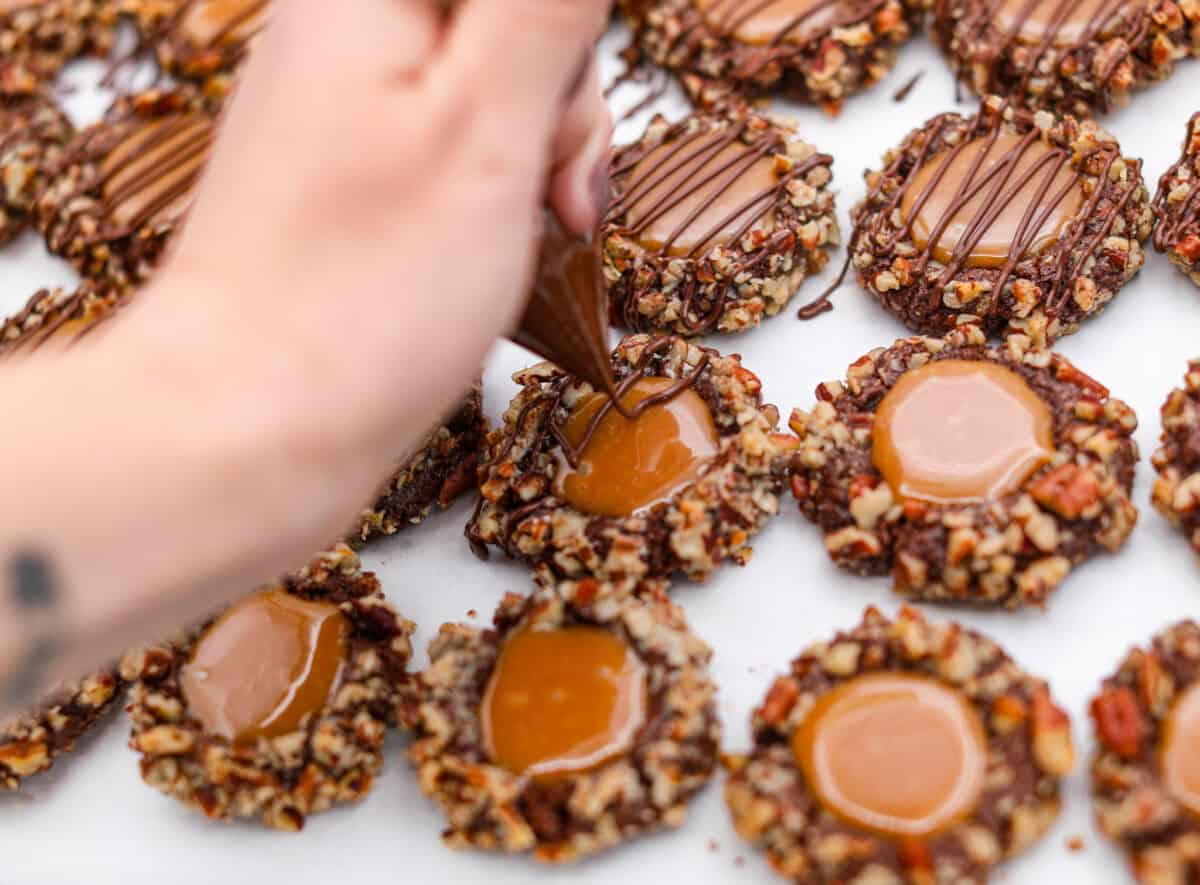 Turtle Cookies are a baked, bite-size brownie loaded with pecans, then filled with a creamy caramel center for a rich, decadent holiday cookie! These turtle cookies are always a huge hit and are gone in minutes Close up shot of someone drizzling chocolate over the turtle cookies.
