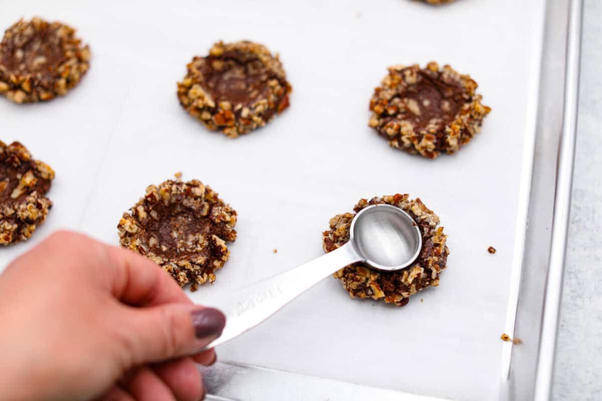 Turtle Cookies are a baked, bite-size brownie loaded with pecans, then filled with a creamy caramel center for a rich, decadent holiday cookie! These turtle cookies are always a huge hit and are gone in minutes Angle shot of someone making indents in the cookie dough balls with pecans.