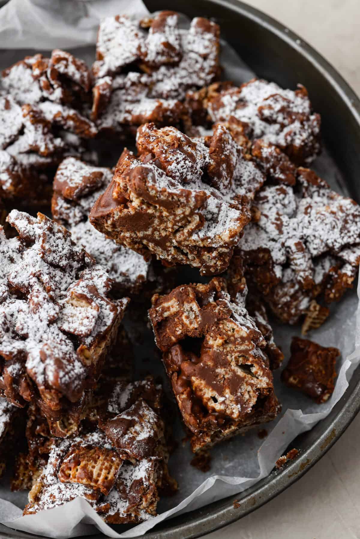 These tasty Muddy Buddy Bars are a fun twist on the classic snack mix. Crunchy cereal is surrounded by sweet marshmallows, chocolate, and peanut butter. These bars are a fun new way to enjoy this classic flavor combination. Close up shot of cut up muddy buddy bars on a serving platter.