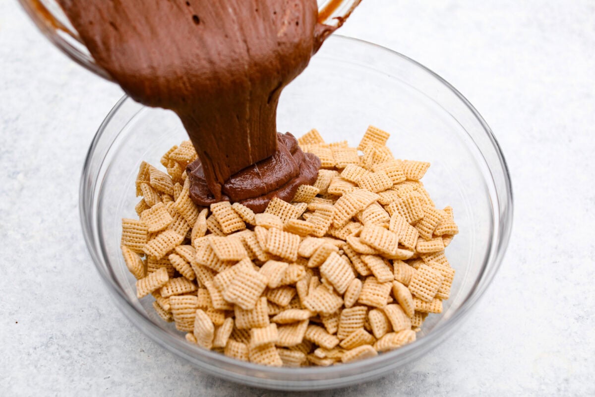 These tasty Muddy Buddy Bars are a fun twist on the classic snack mix. Crunchy cereal is surrounded by sweet marshmallows, chocolate, and peanut butter. These bars are a fun new way to enjoy this classic flavor combination. Overhead shot of someone pouring the chocolate and marshmallow mixture over the cereal.