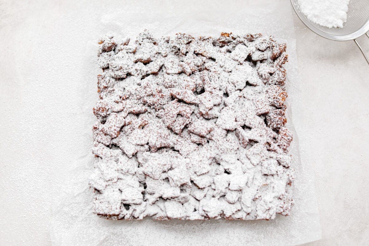 These tasty Muddy Buddy Bars are a fun twist on the classic snack mix. Crunchy cereal is surrounded by sweet marshmallows, chocolate, and peanut butter. These bars are a fun new way to enjoy this classic flavor combination. Overhead shot of muddy buddy bars dusted with powdered sugar.