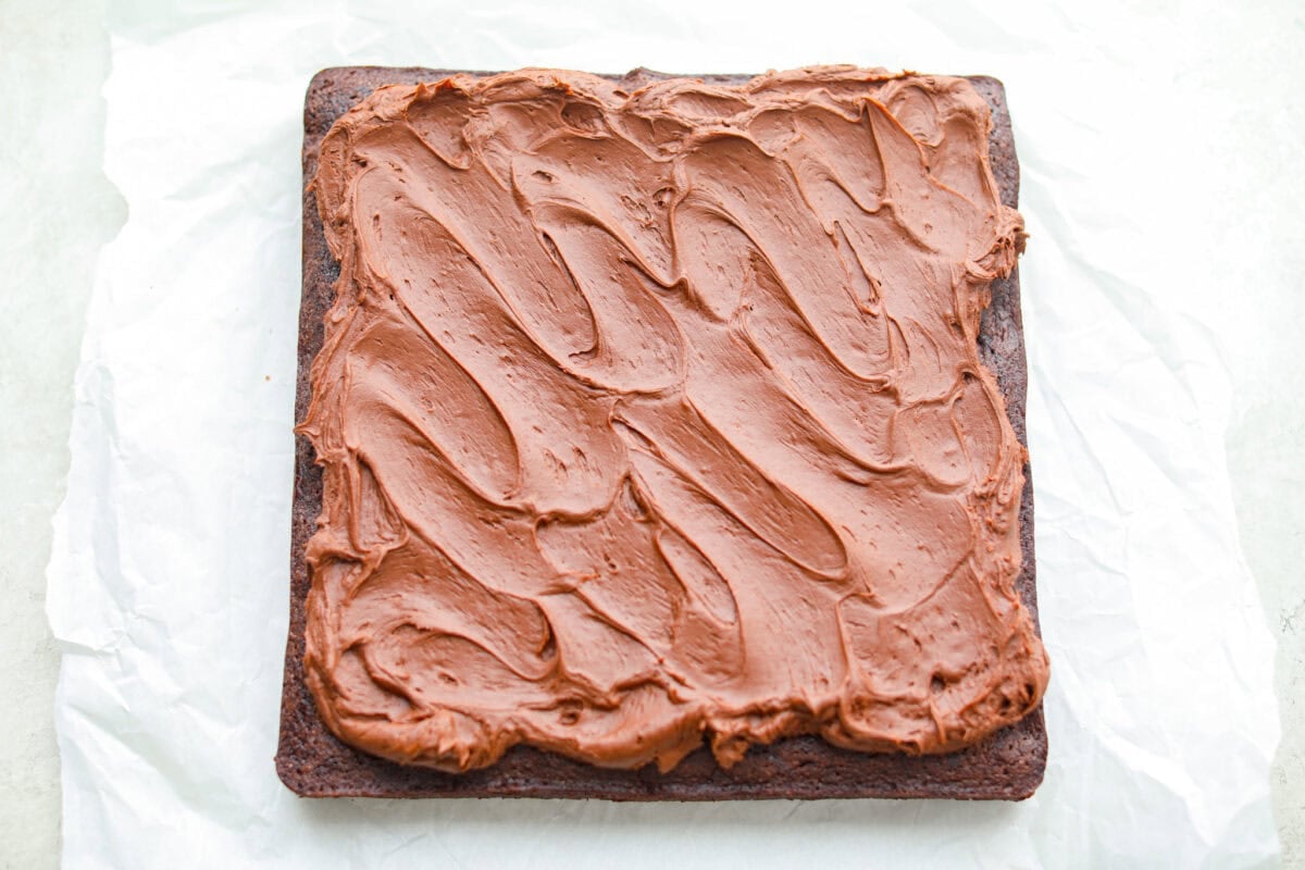 Rich, fudgy, and packed with deep chocolate flavor, Guinness Brownies are a must-try for any dessert lover! The stout beer enhances the cocoa, adding a subtle, malty depth of flavor that makes these brownies rich and irresistibly delicious. Overhead shot of baked and frosted Guinness brownies on a piece of parchment paper.